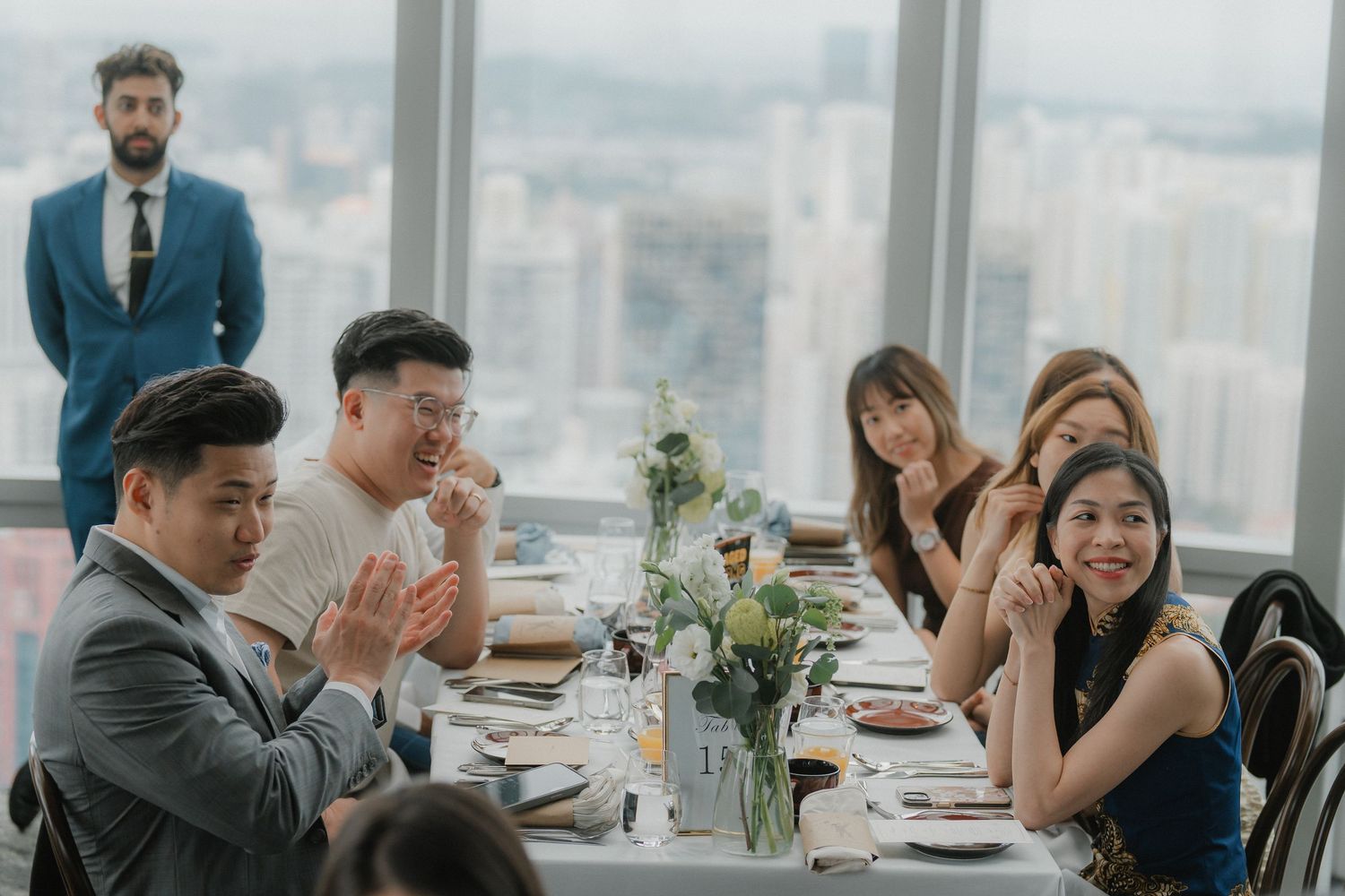 Wedding reception guests seated at long tables in a modern venue with panoramic city views.