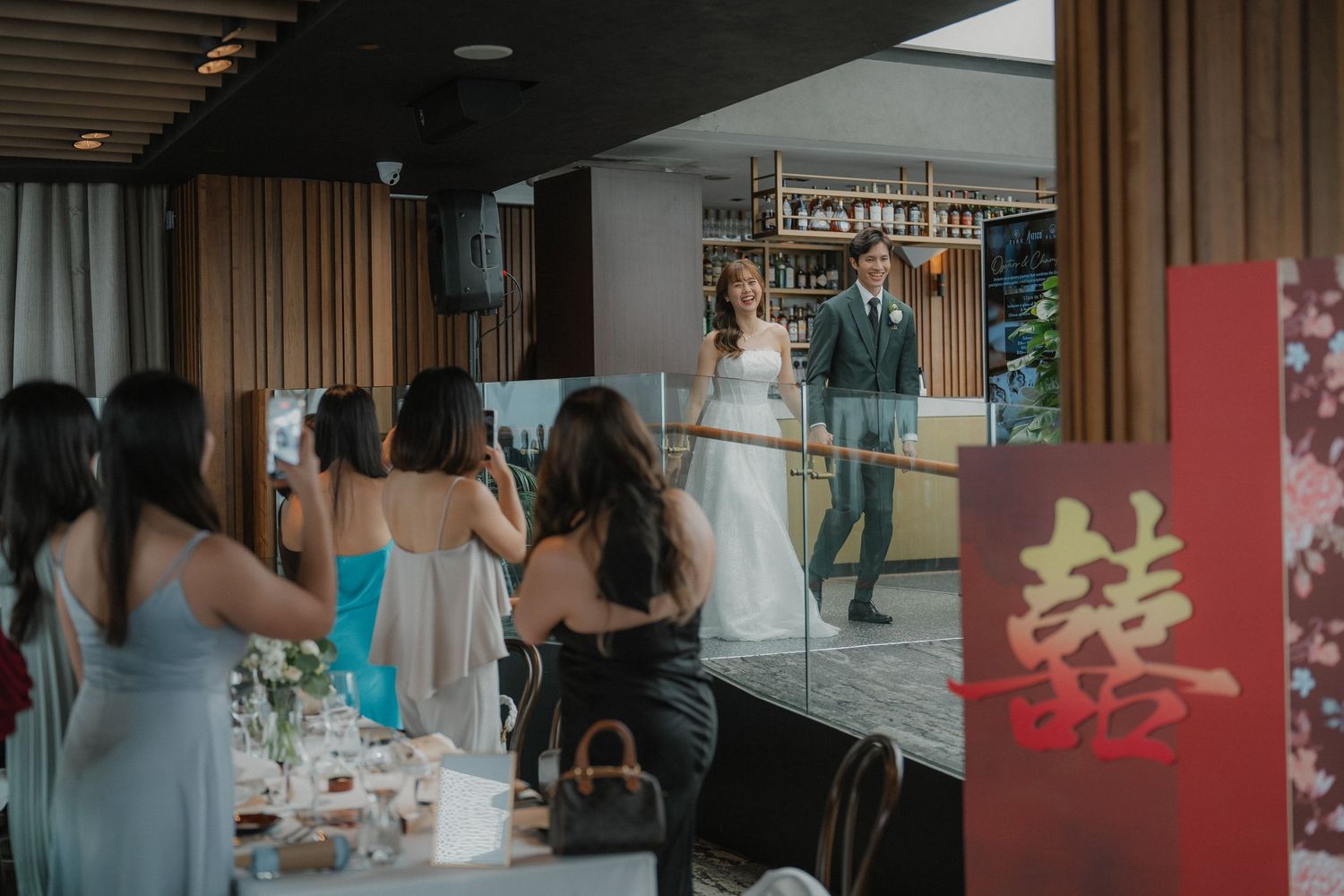 Wedding guests at a modern reception venue enjoy a celebration with white curtains and wooden accents in the background.