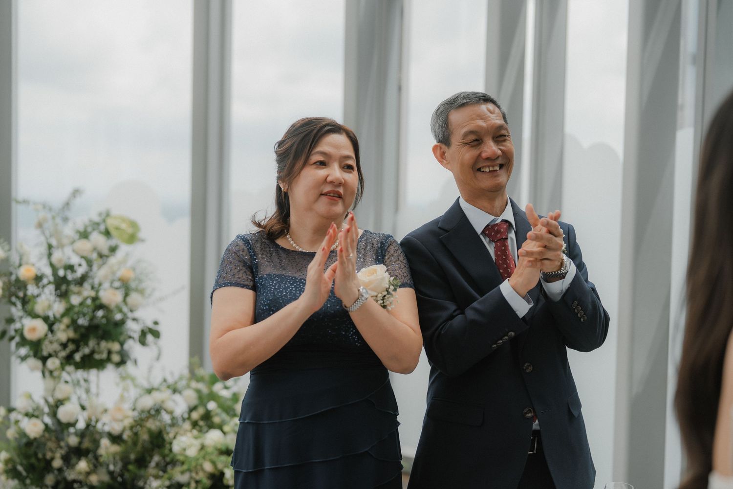 Wedding guests clap and smile during a joyous celebration in front of white floral decorations.