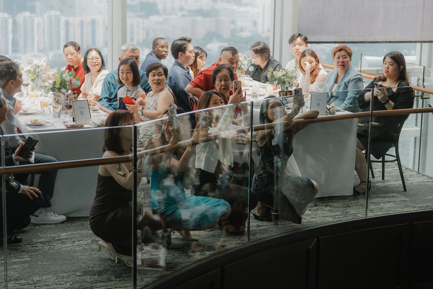Wedding celebration with guests seated at long tables beside large windows with city views.