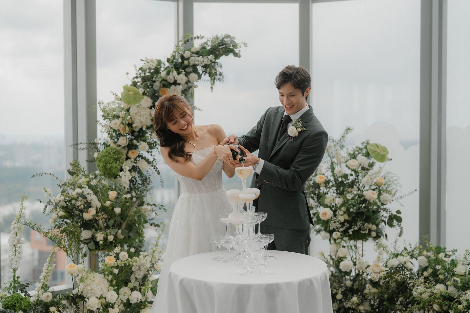 Newlyweds cutting cake together by floral arch near floor-to-ceiling windows with city views.