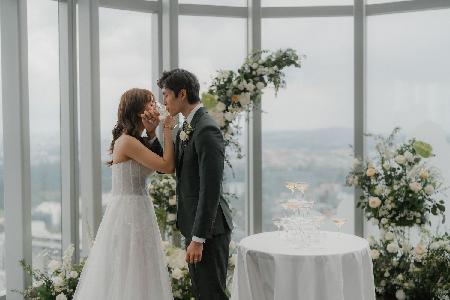A romantic wedding moment in a bright windowed venue overlooking a city skyline with floral decor.