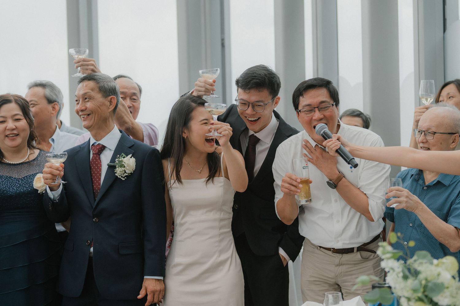 Wedding celebration with guests toasting and cheering in front of large windows.
