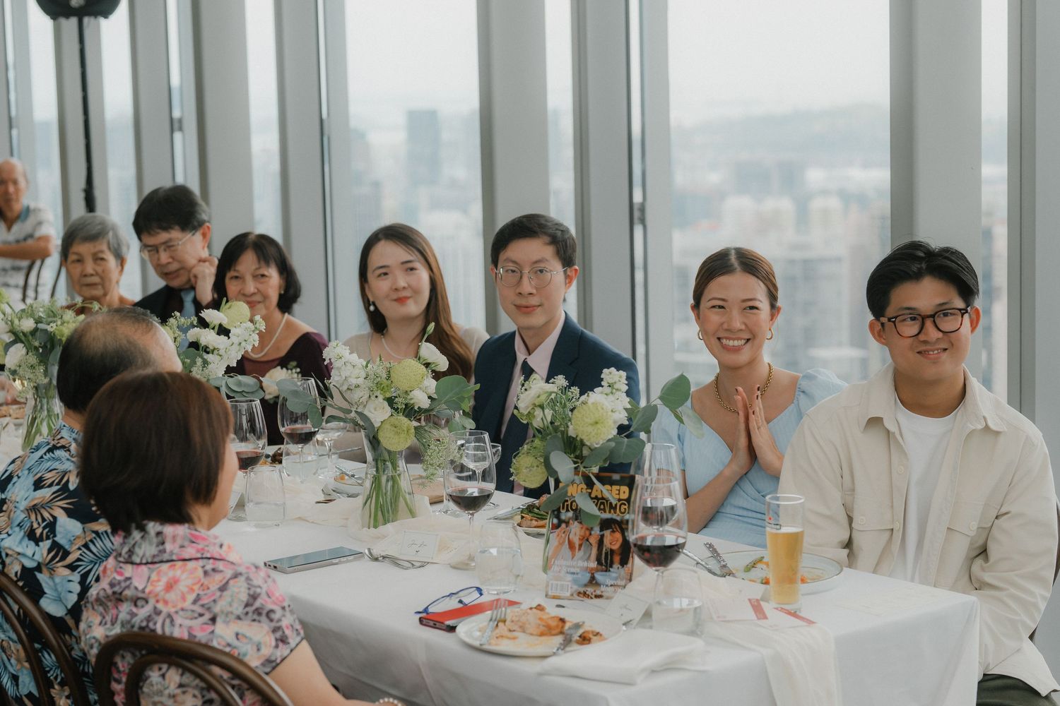 Wedding reception guests dining at round table with white floral centerpieces and city views.
