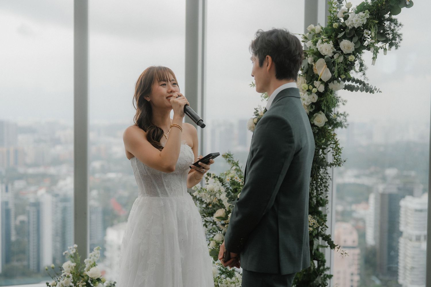 Couple exchanging vows at rooftop wedding venue with cityscape backdrop and floral decorations.