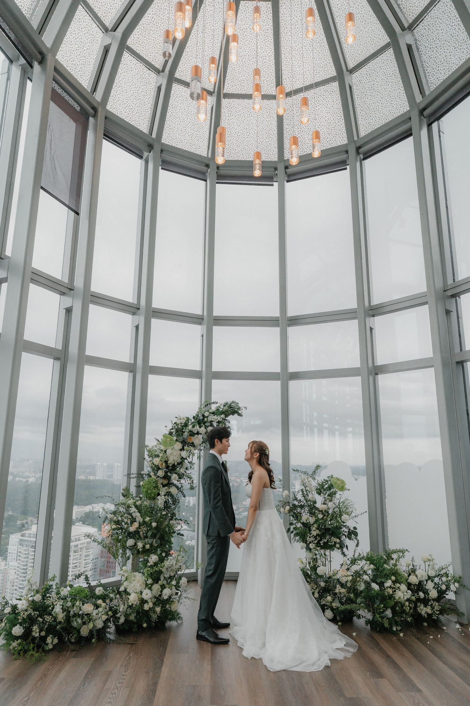 Elegant wedding couple standing in front of floral arrangements in a glass-domed room overlooking the city.