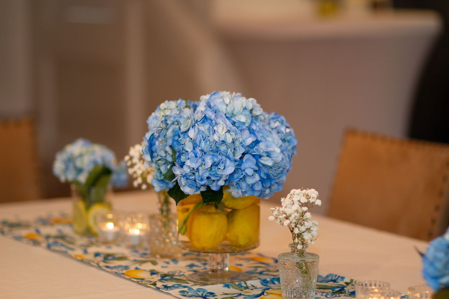 Elegant table settings feature blue hydrangeas and white baby's breath centerpieces with glowing candles and floral patterns.