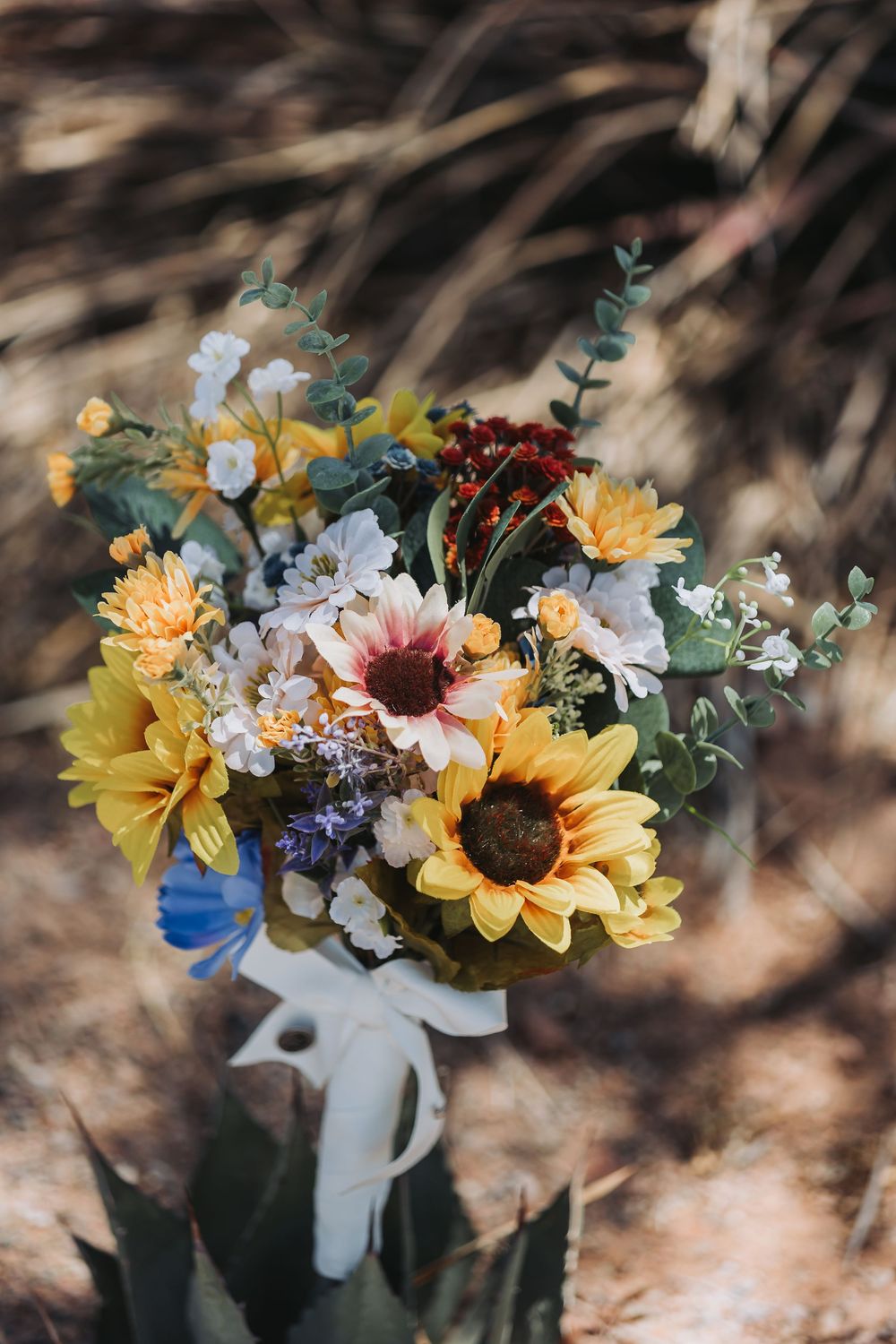 A rustic bouquet featuring sunflowers, pink and yellow blooms with eucalyptus leaves tied together with a white ribbon.