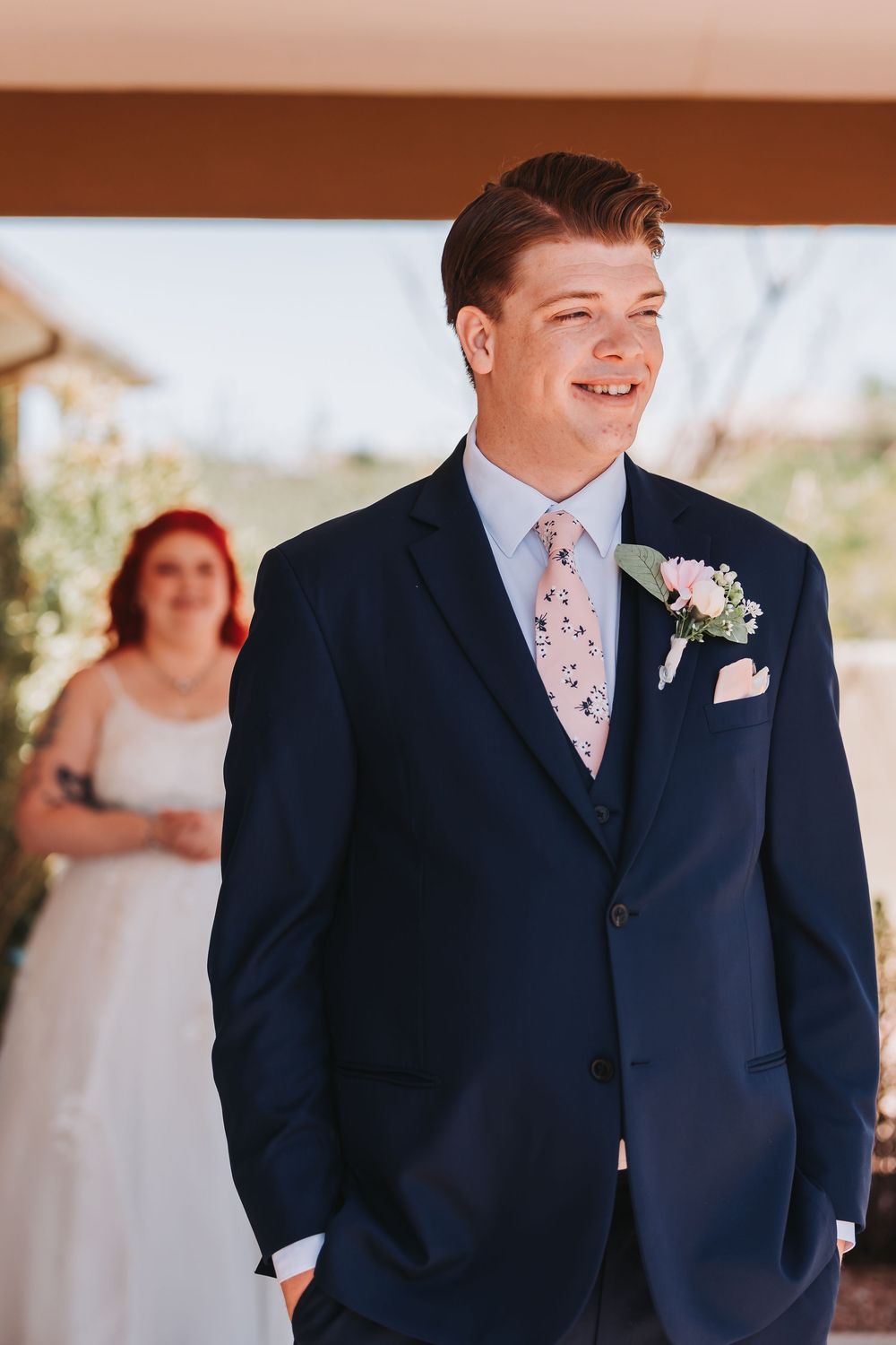 The groom in a navy suit with floral tie smiles joyfully during an outdoor wedding ceremony.