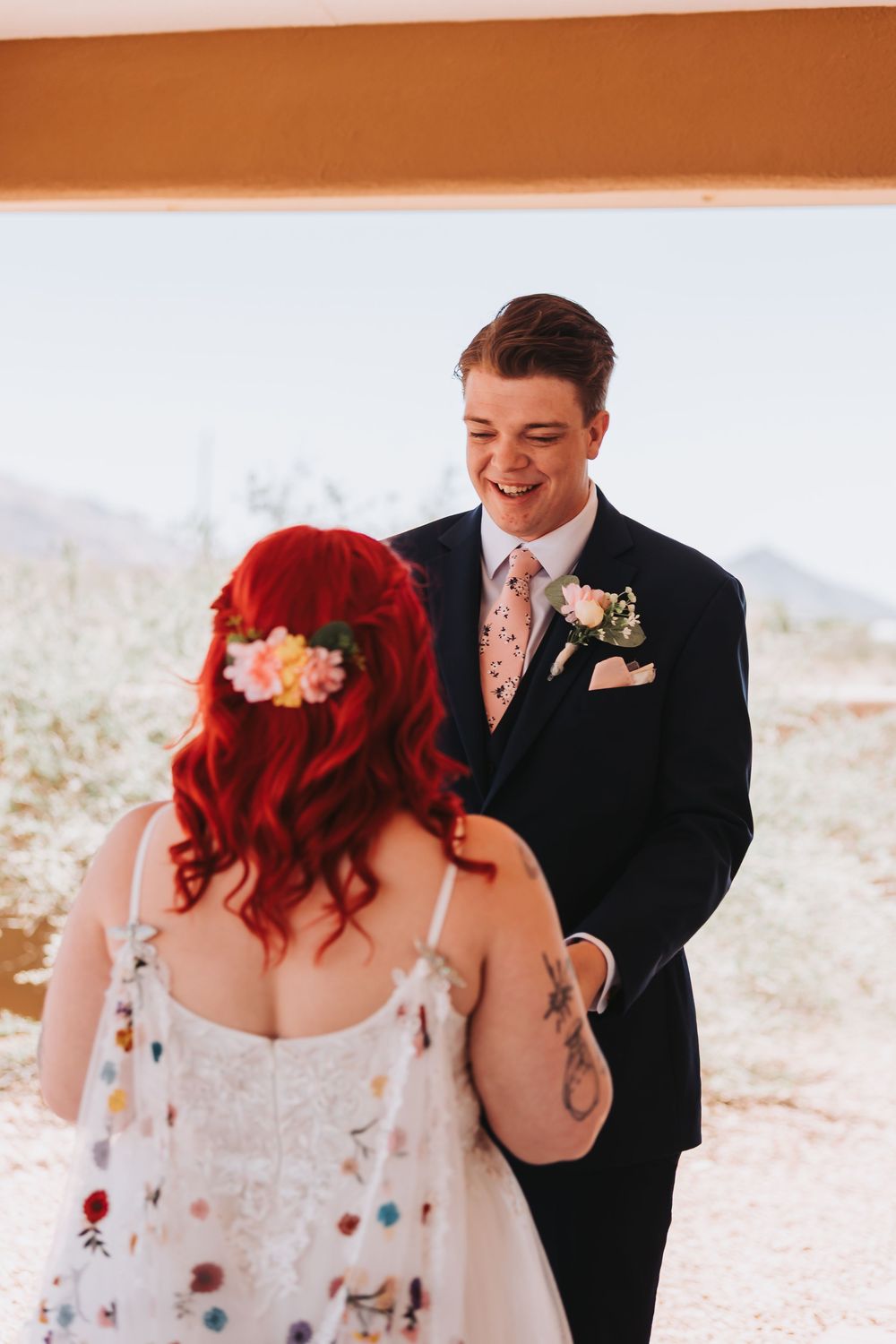 A wedding first look moment shows a groom in black suit reacting with joy at seeing the bride's red hair and white dress.