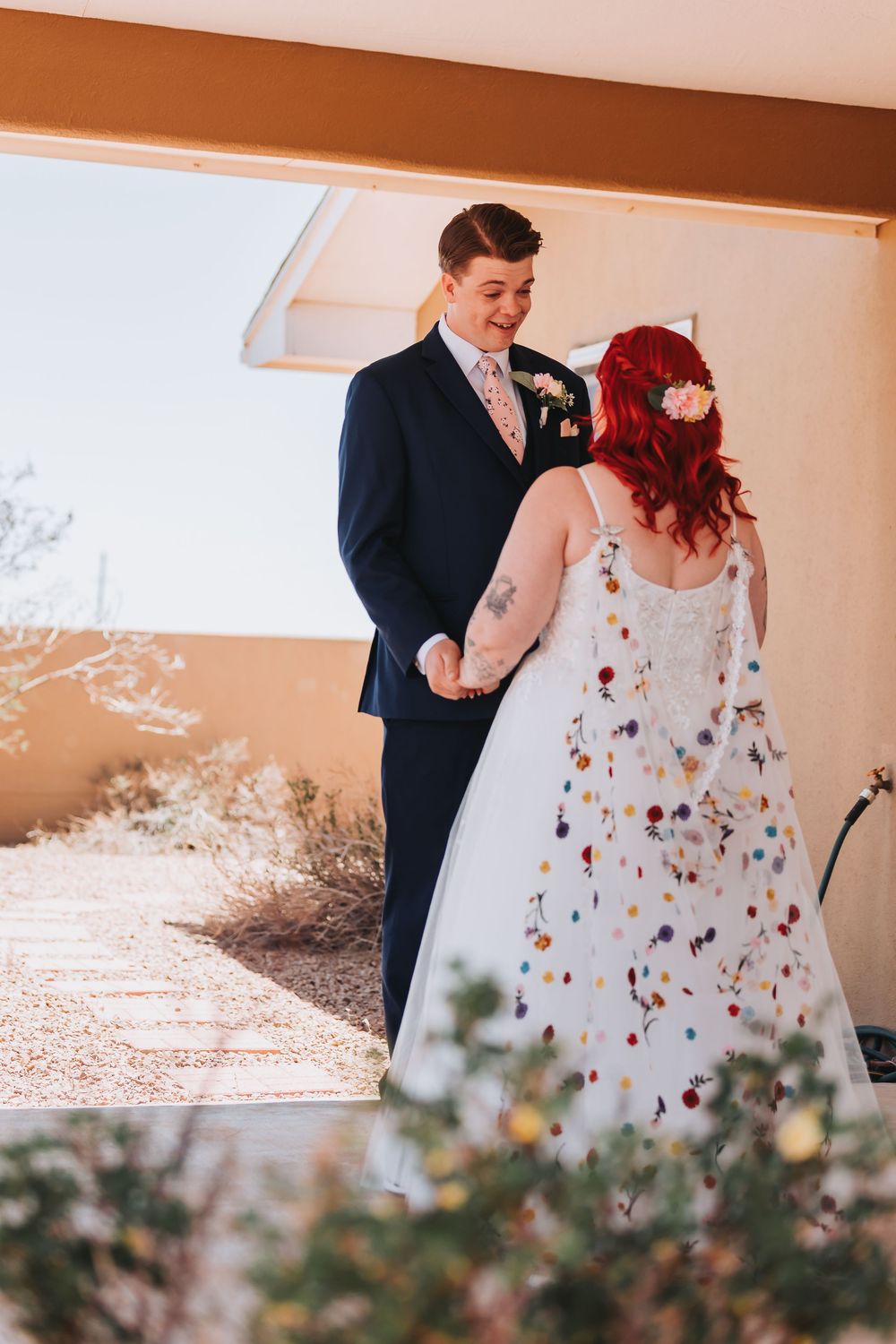 Bride in a floral adorned white dress stands with her partner in a navy suit during an outdoor desert wedding ceremony.