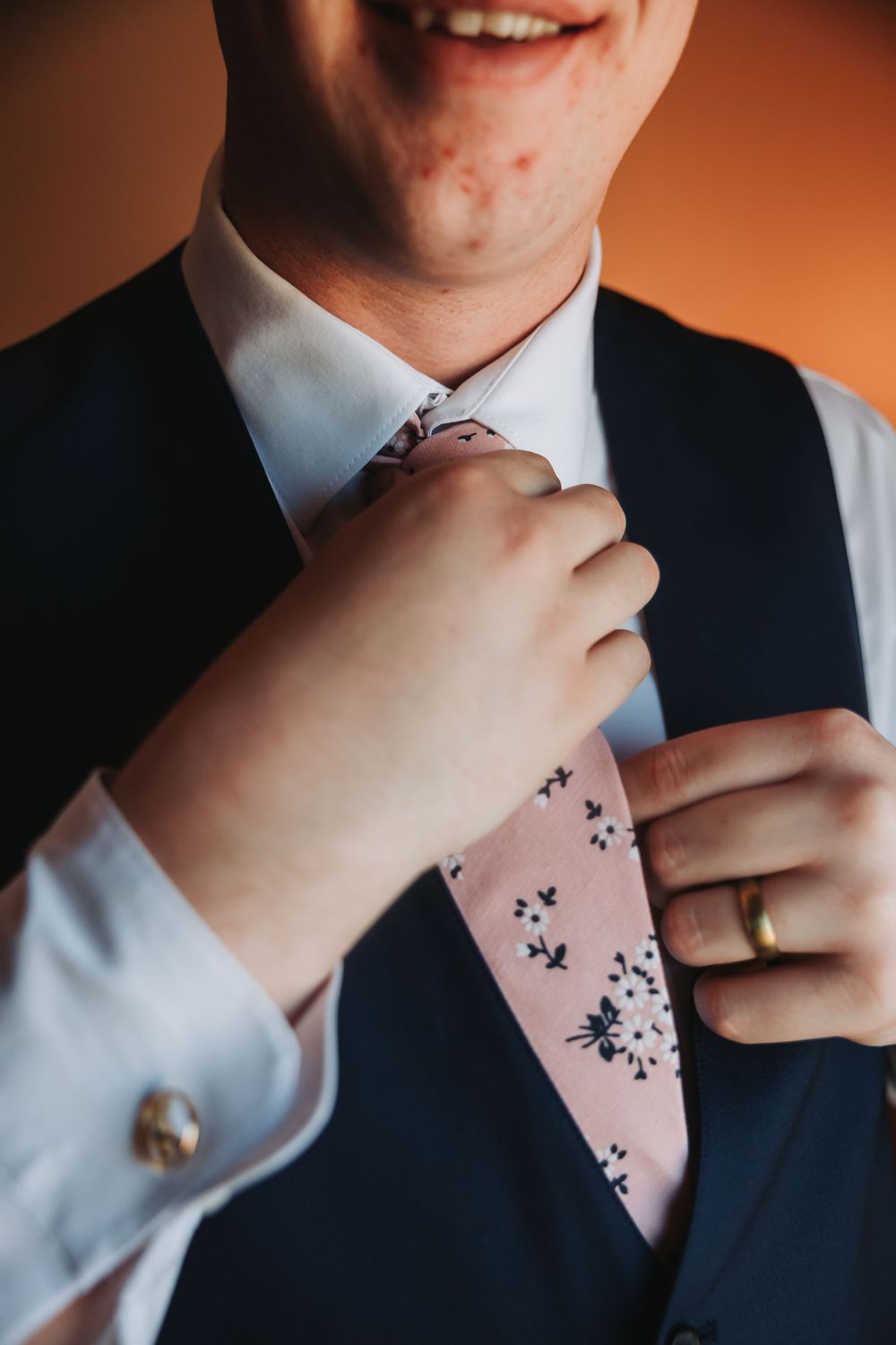 Close-up shot of a person adjusting a pink floral tie while wearing a navy vest and white dress shirt with a gold ring.