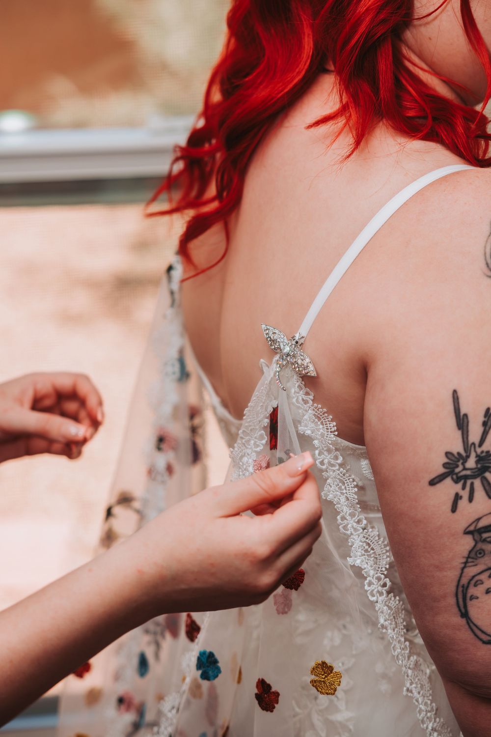 Hands adjusting delicate lace straps on a white dress while vibrant red hair cascades over shoulder with visible tattoo.