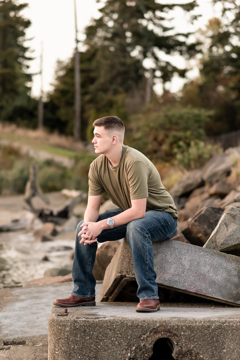 Someone sits thoughtfully on concrete blocks along a rocky shoreline wearing a green t-shirt and jeans at sunset.