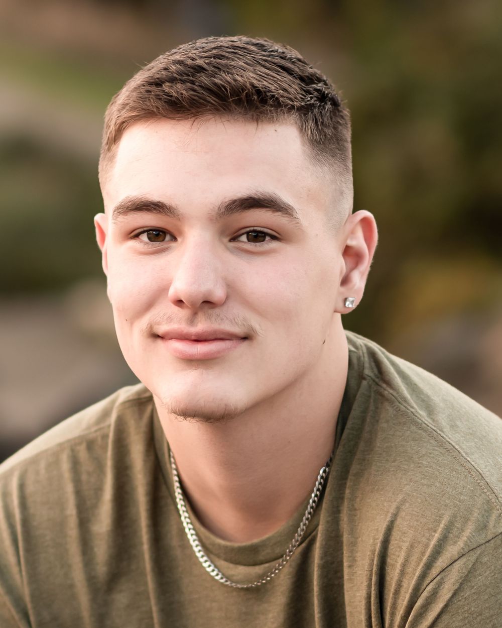 Young person smiling in a casual olive green t-shirt with a silver chain necklace against a blurred outdoor background.