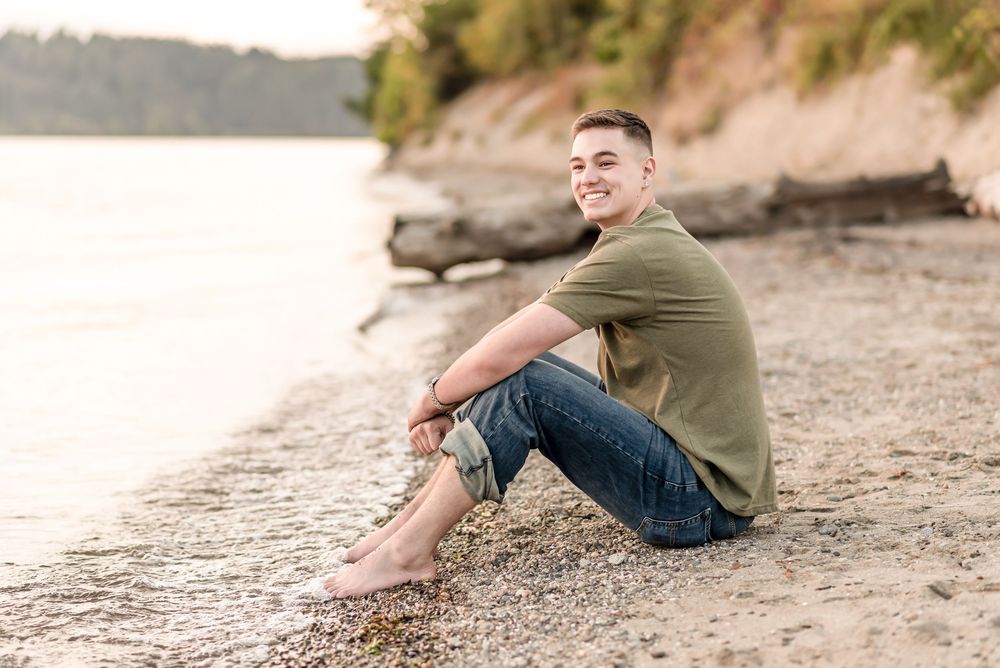 A thoughtful person sits barefoot on a sandy beach looking out at the ocean at sunset.