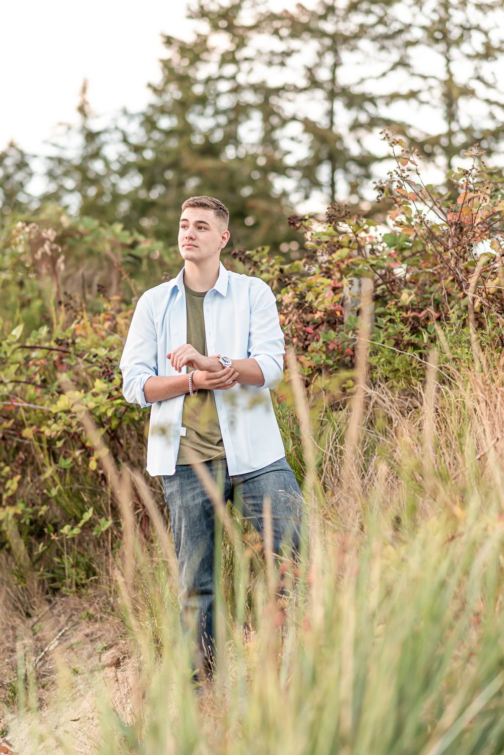 Someone poses outdoors in casual attire against a backdrop of autumn foliage and evergreen trees.