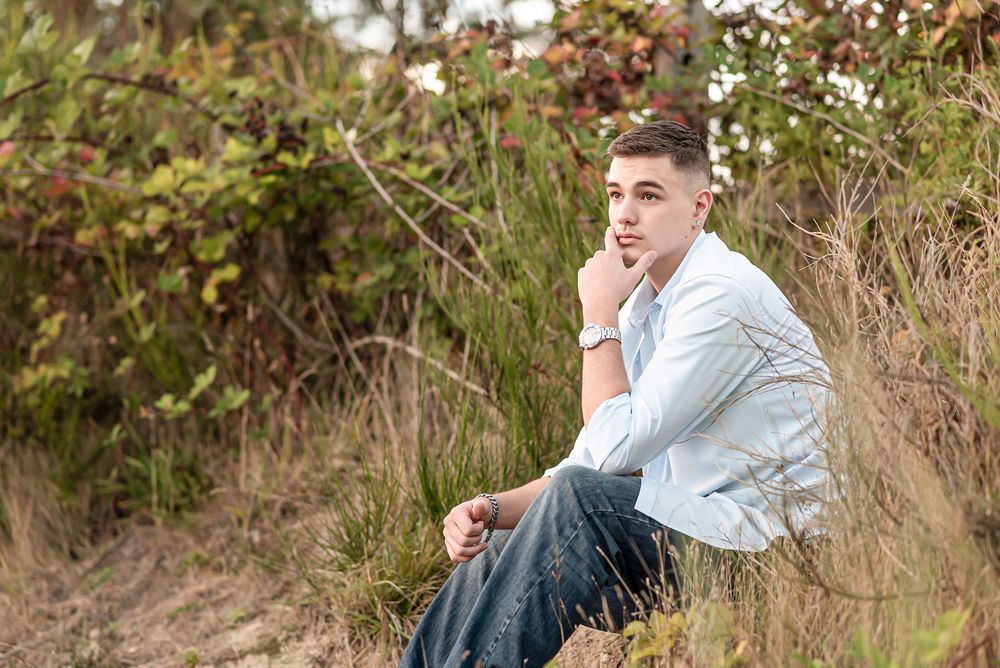 Someone wearing a light blue shirt and jeans sits thoughtfully in a natural outdoor setting with flowering bushes.