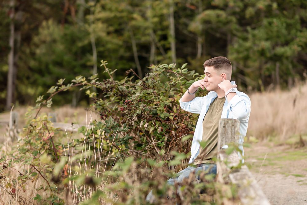 A person stands along a forest path, wearing casual clothing and looking off into the distance on a sunny day.