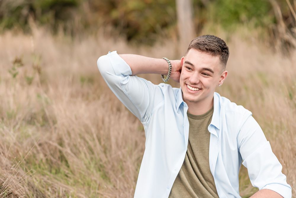 A person wearing a white and olive shirt smiles and poses playfully in a grassy outdoor setting.