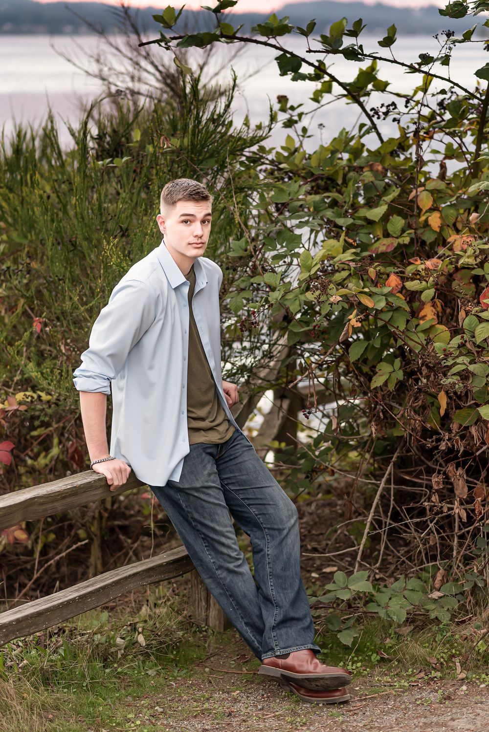 Casual portrait in nature showing person leaning against wooden railing wearing light button-down shirt and jeans.