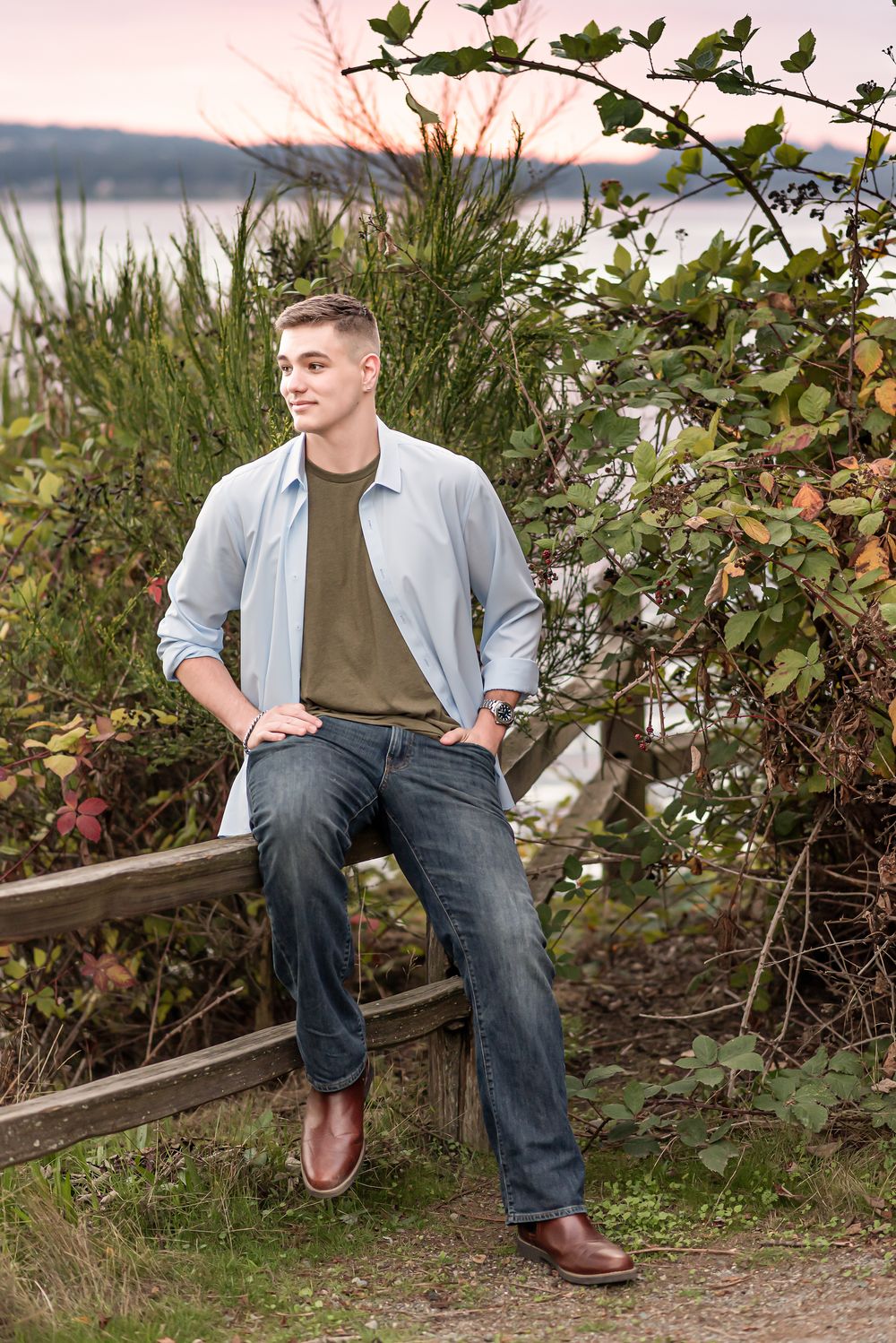 A person in casual attire sits on a wooden bench surrounded by lush greenery near a waterfront at sunset.