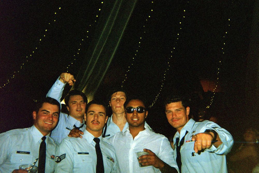A group of people wearing military uniforms laugh and pose together at night with laser beams visible in background.