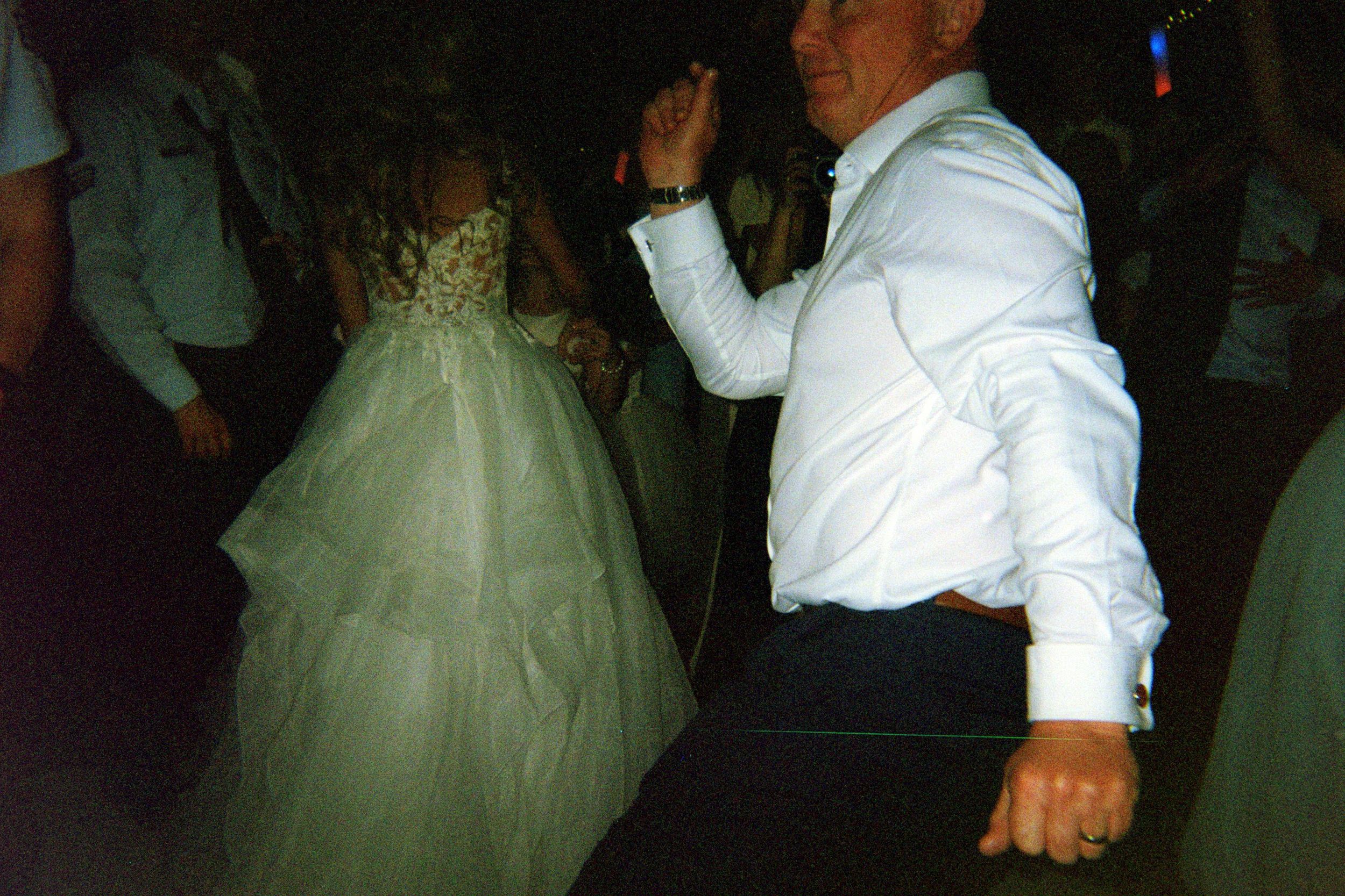 Two people in wedding attire dance together at a reception while guests look on.