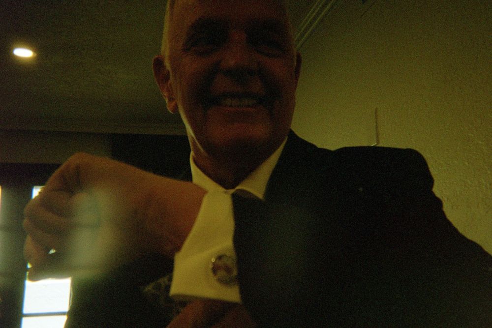 A person in a dark suit smiling in dim lighting conditions with a yellowish ambient glow.