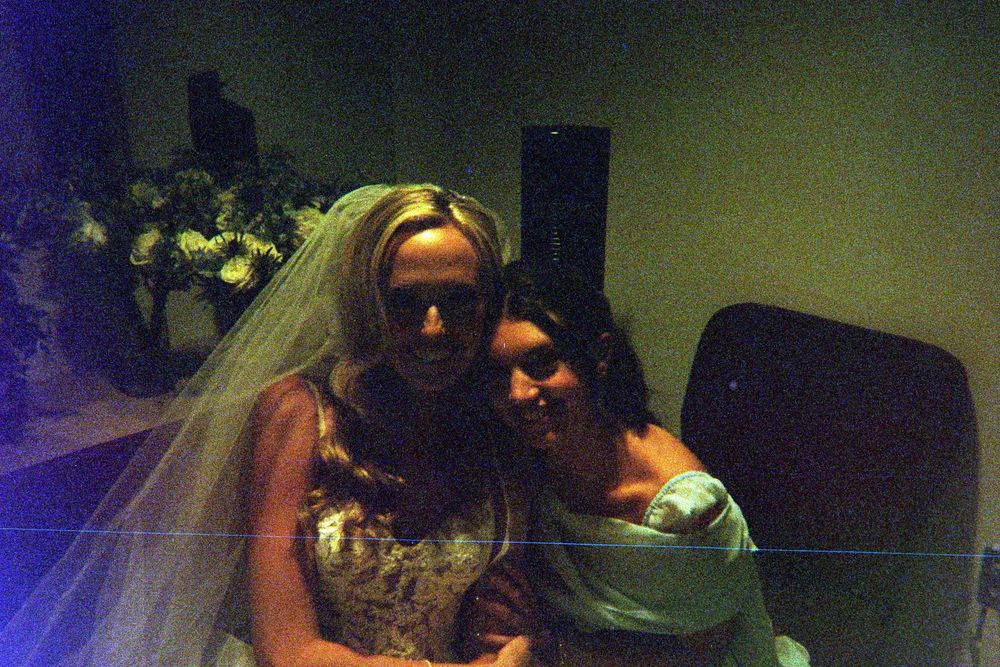 A bride and companion pose together at a dimly lit wedding reception with teal lighting and floral decorations in background.