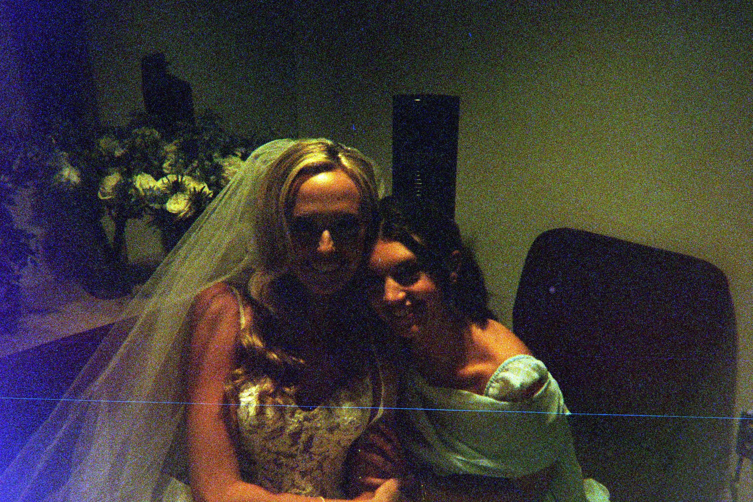 A bride and companion pose together at a dimly lit wedding reception with teal lighting and floral decorations in background.