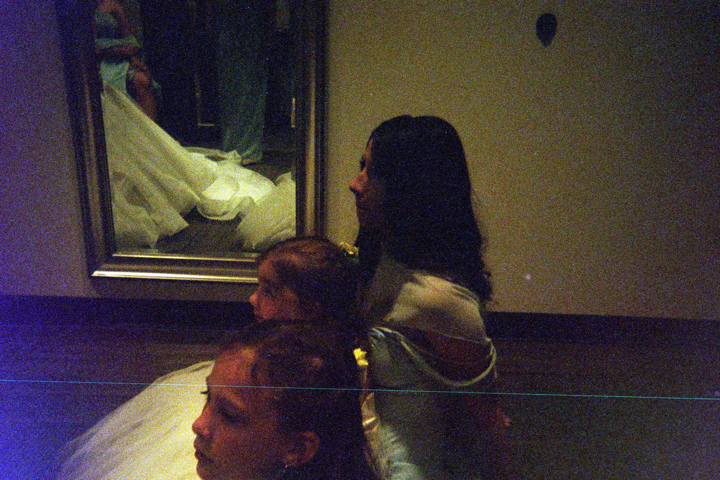 A figure in a white dress is reflected in a mirror in a dimly lit room with purple lighting.