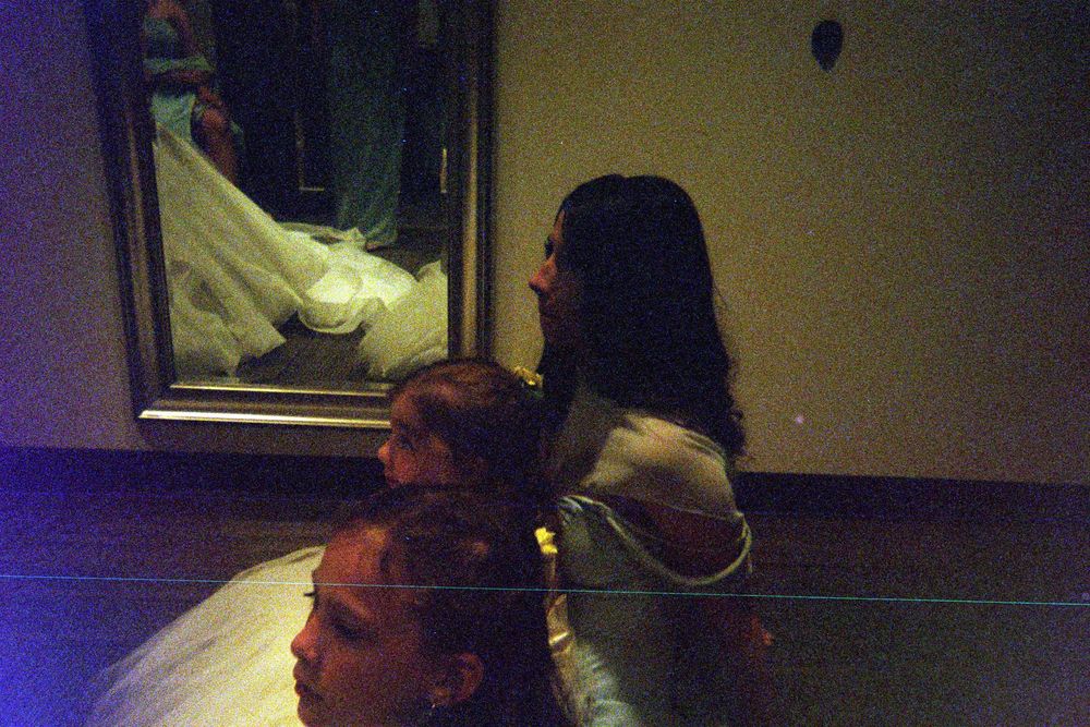 A figure in a white dress is reflected in a mirror in a dimly lit room with purple lighting.