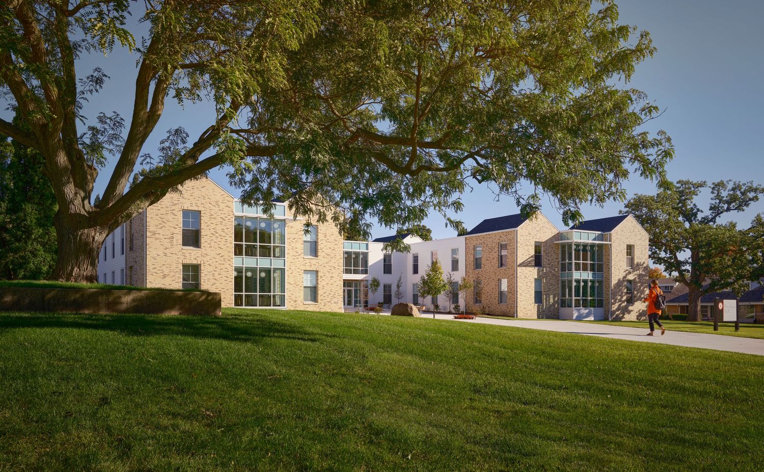 Modern residential buildings with large windows sit on a green lawn under mature trees at sunset.