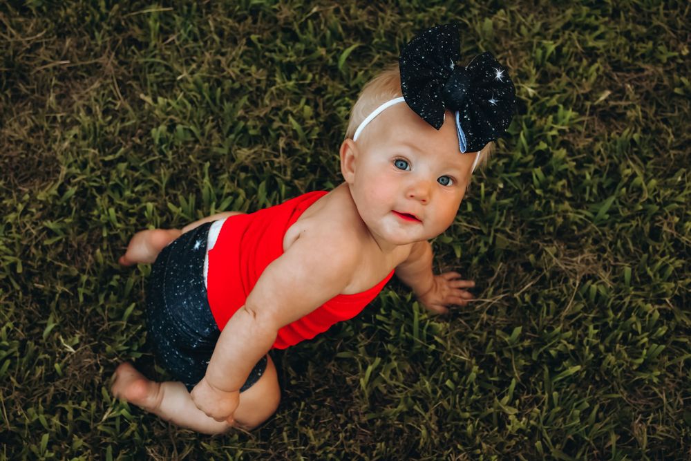 A baby wearing a red romper lies on grass outdoors on a sunny day.