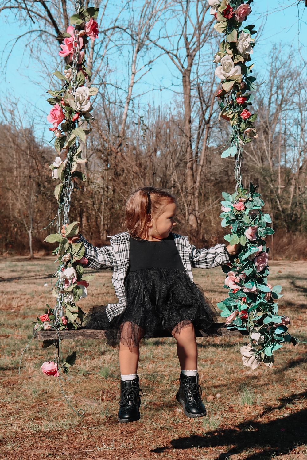 A child in a black dress and boots sits on a floral decorated swing in a wooded outdoor setting during autumn.
