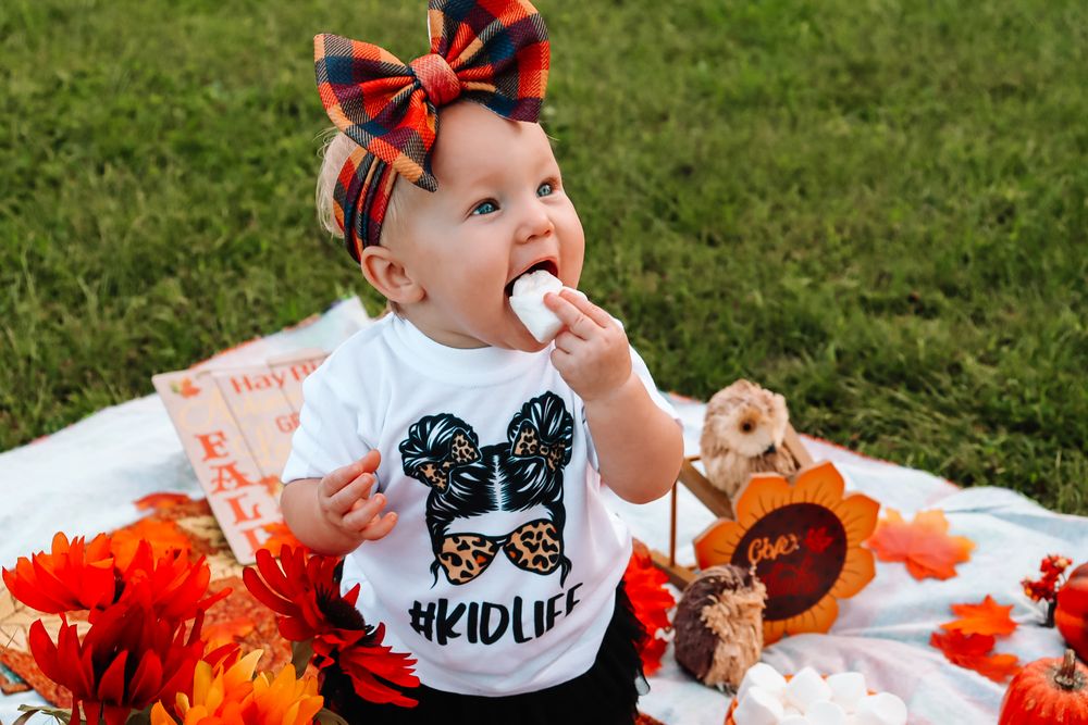 A baby in a butterfly shirt sits on a fall-themed picnic blanket with orange decorations and a plaid bow.