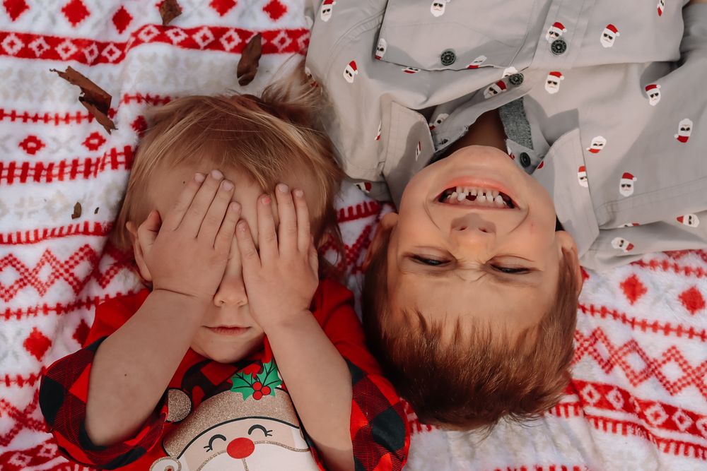 Two children laying on red and white holiday patterned bedding playfully express joy during Christmas season.
