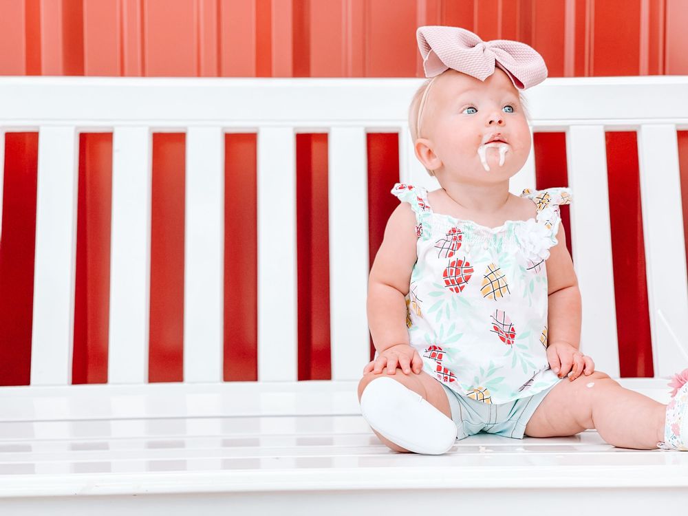 Baby in floral romper sits on white bench against red and white striped background.