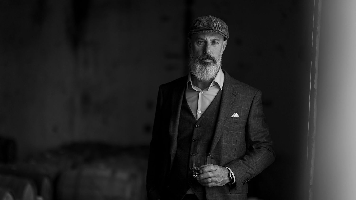 Black and white portrait of a bearded gentleman in vintage suit, flat cap and vest holding a drink at Corowa Whisky.
