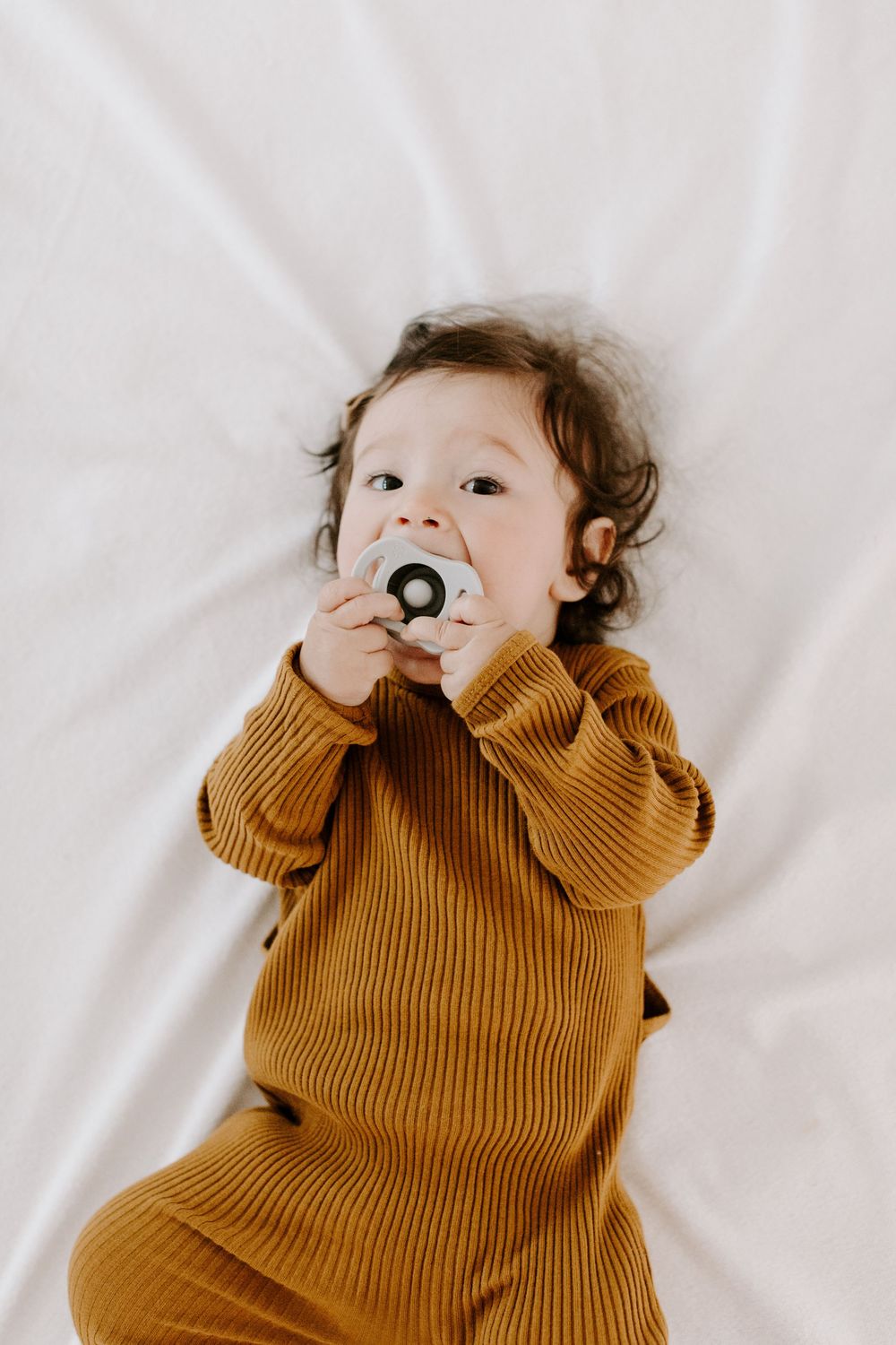 A small child in a cozy mustard yellow ribbed outfit lies on a white background while holding a pacifier.