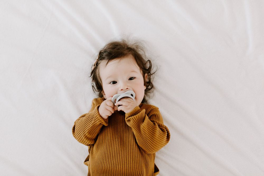 Baby wearing brown knit sweater lying on white blanket while sucking on fingers.