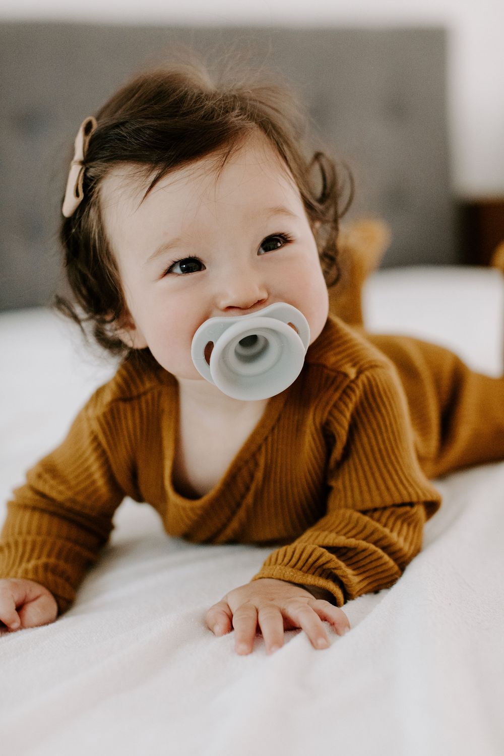 Baby wearing brown sweater lies on white bedding while using a gray pacifier.