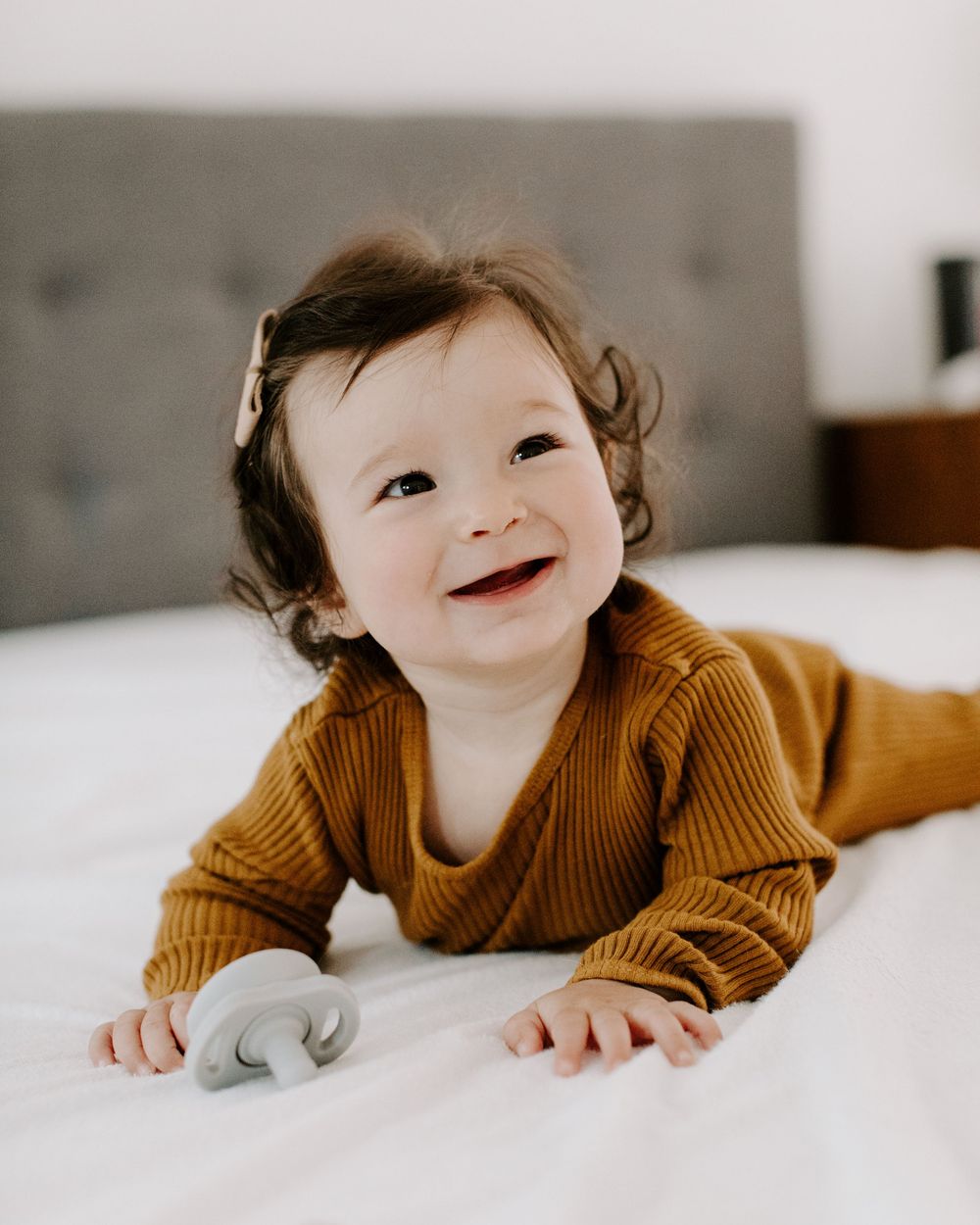 A baby in a brown sweater lies on crisp white bedding with a joyful expression looking upward.