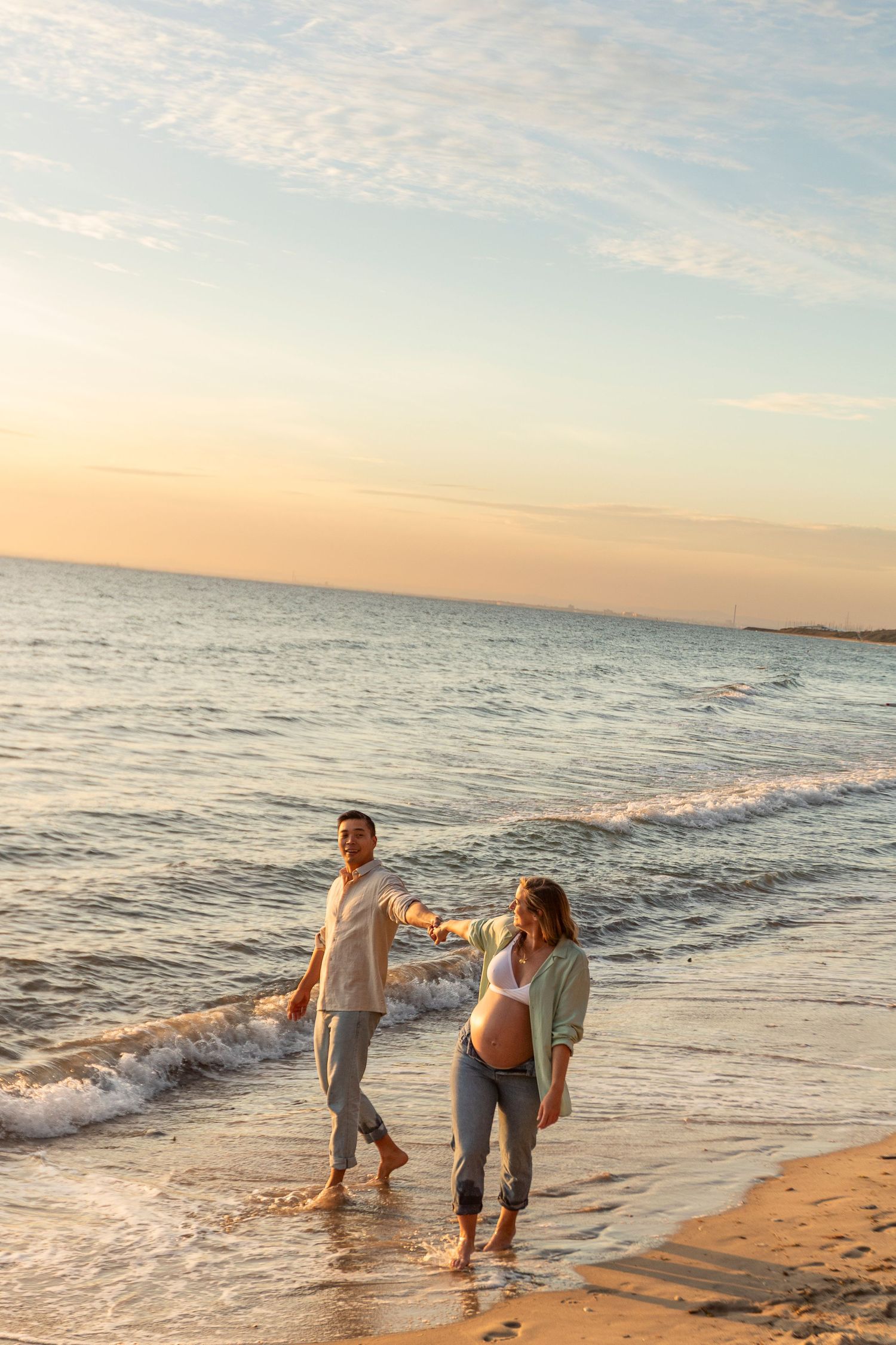 A couple walks along the beach at sunset with waves gently rolling in for their maternity session
