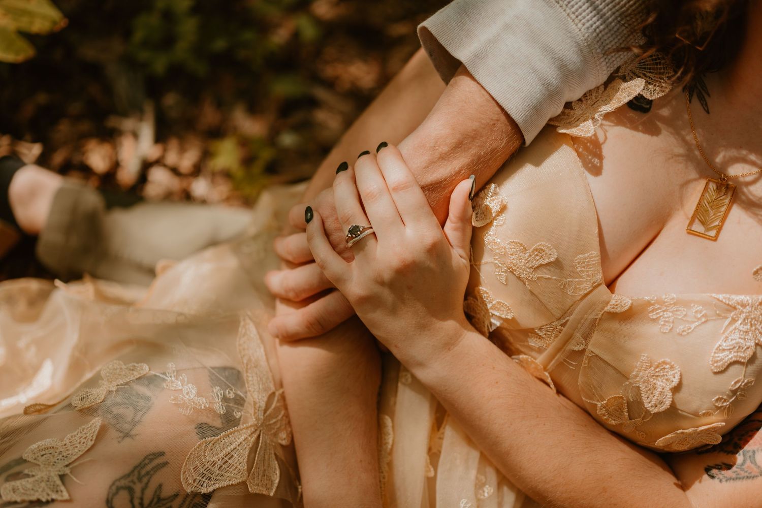 Closeup of a tattooed bride and groom, the bride leaning against her partner, showing off her moss agate engagement ring