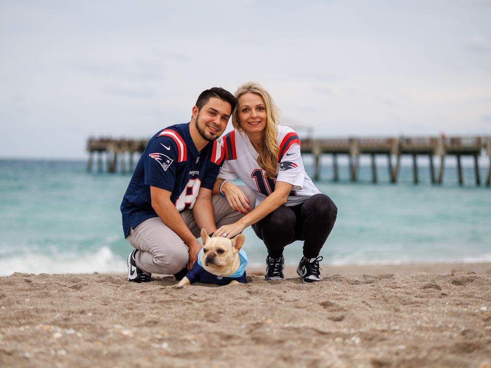 A couple and their dog pose together on a sunny beach with a pier in the background.