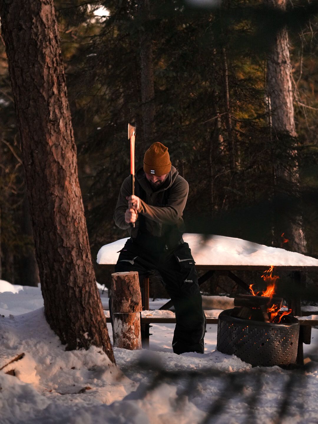 Outdoor winter camping scene with a campfire glowing between snow-covered trees at dusk.