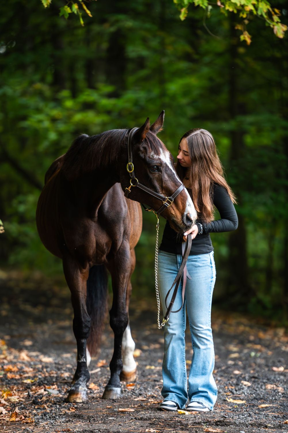 A dark brown horse and rider share a tender moment on an autumn trail surrounded by lush green foliage.