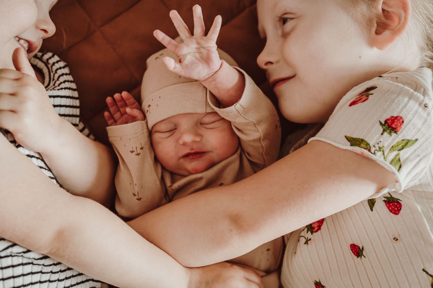 Siblings cuddling with newborn baby on bed during lifestyle photoshoot.